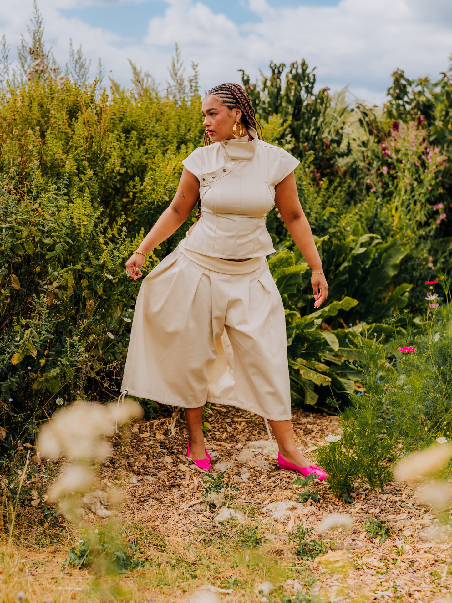 A woman wearing a cream-colored two-piece matching set consisting of a short-sleeve top and high-waisted pleated skirt, standing outdoors in a garden setting with green foliage and dry grass.
