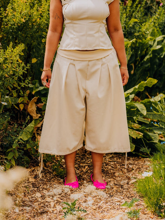 A woman stands outdoors in a garden wearing a light beige two-piece outfit consisting of a short-sleeve wrap-style top and matching wide-leg cropped pants, paired with bright pink shoes.