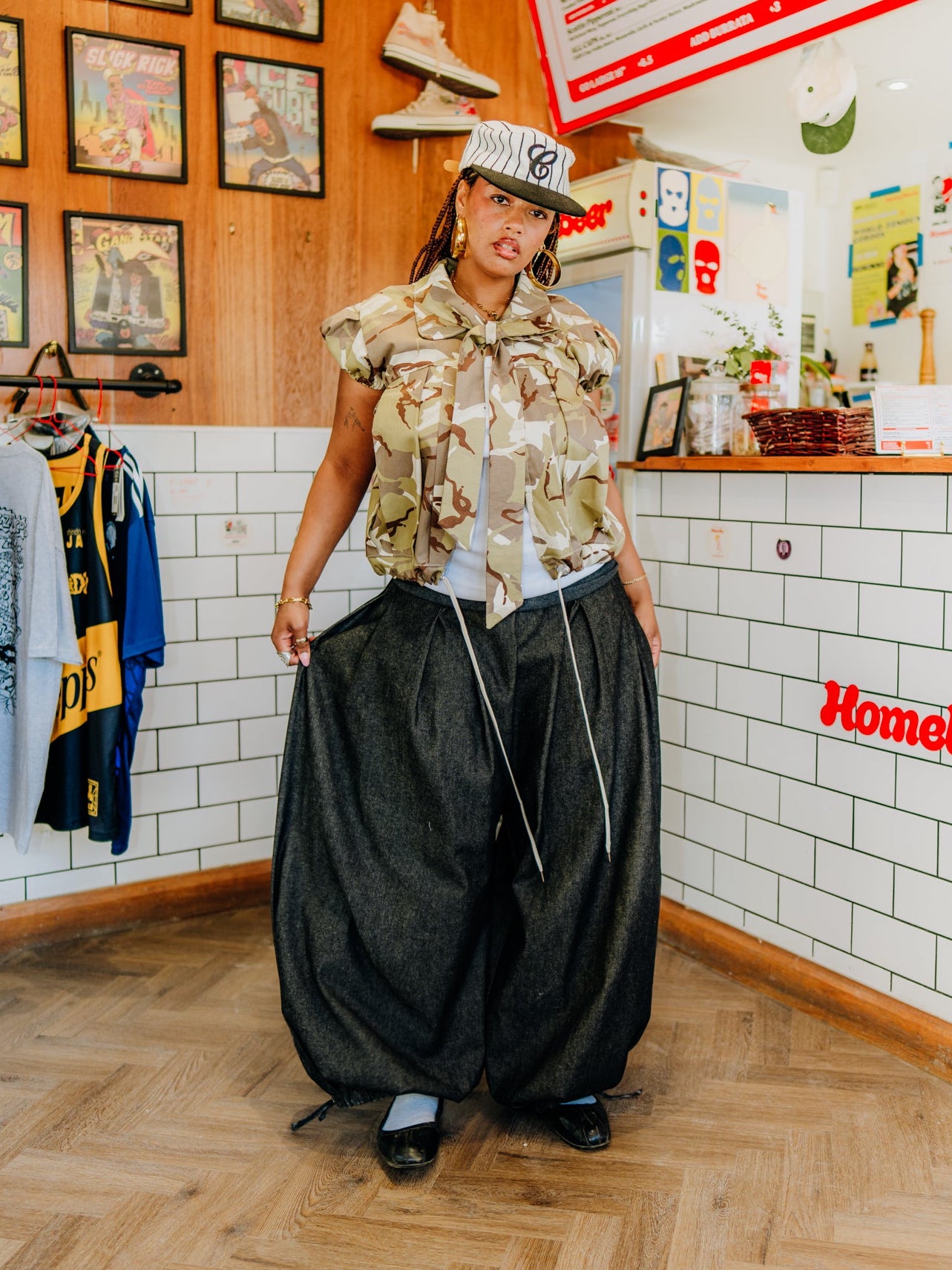 A person stands in a retail store wearing a beige camouflage patterned short-sleeve shirt, oversized dark gray baggy pants, and a white cap with a black logo. The background features a wood-paneled wall with framed pictures, a clothing rack, and a white tiled counter with a red 'Homeboy' sign.